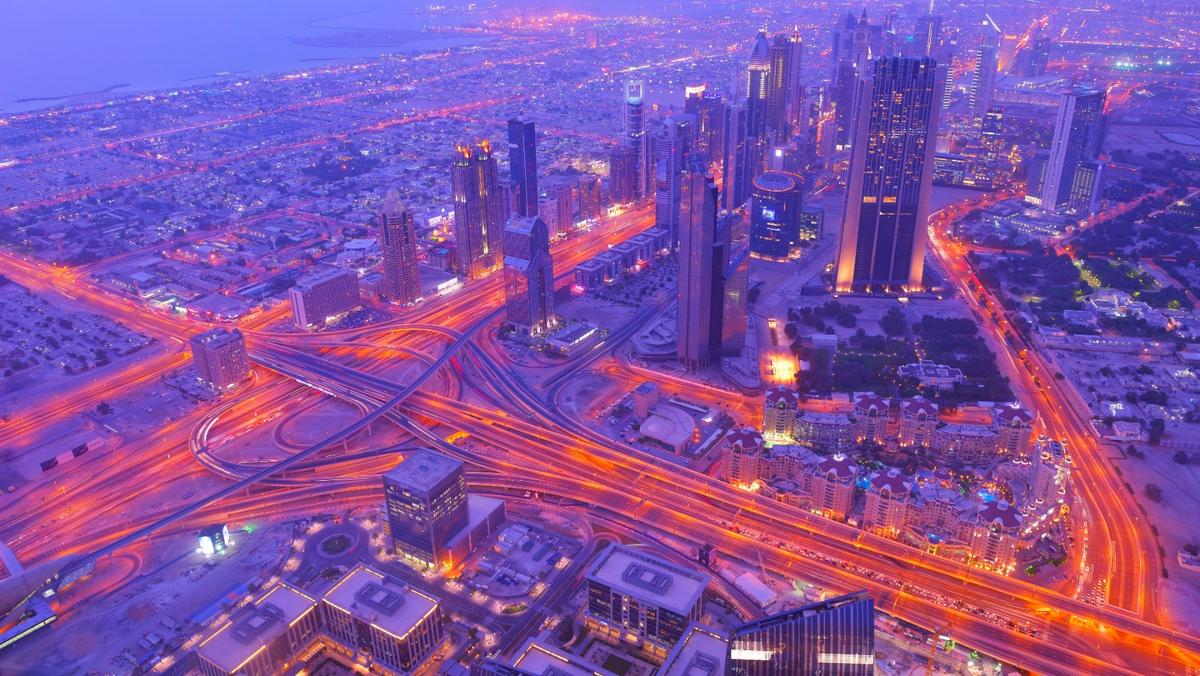 Aerial view of Dubai's skyline and glowing highway interchanges at twilight, bathed in purple and orange light.
