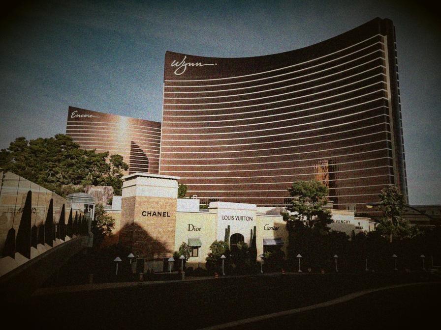 The curved bronze towers of the Wynn and Encore hotels in Las Vegas, with luxury storefronts like Chanel and Dior in the foreground.