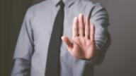 A person in a grey dress shirt and tie holding up an open palm in a stop gesture.
