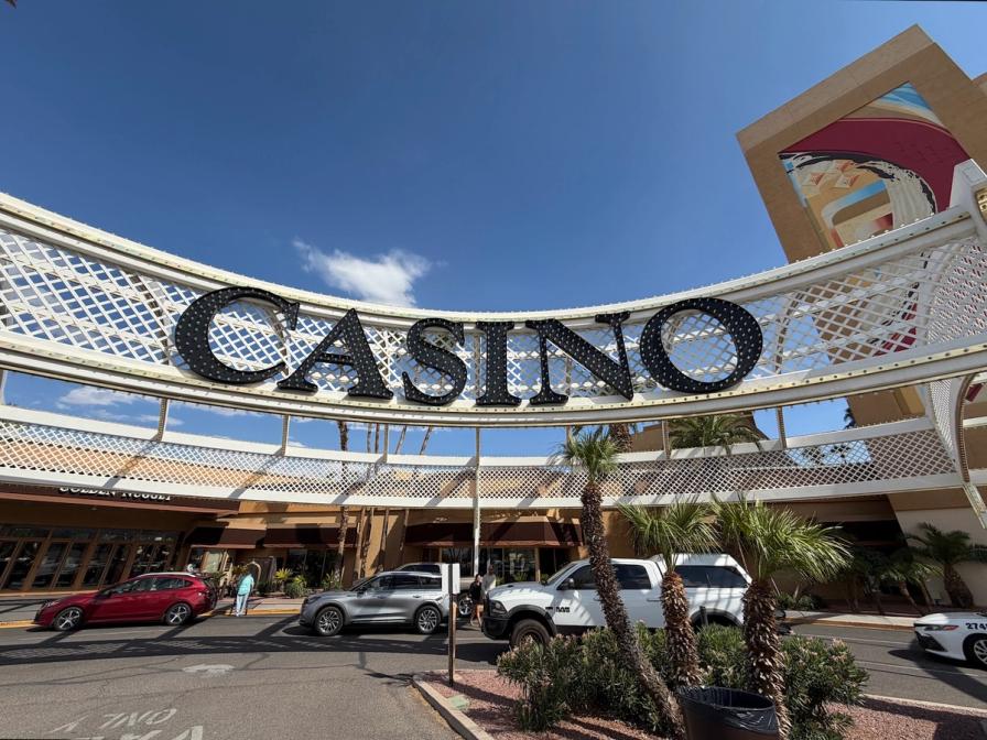 A large white archway with a black CASINO marquee sign over a driveway with cars and palm trees under a clear blue sky.
