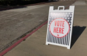 A white A-frame sign on a sidewalk with a patriotic design reads "VOTE