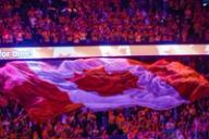 A large Canadian flag is unfurled over a crowd of Calgary Flames hockey fans wearing red jerseys in an arena.