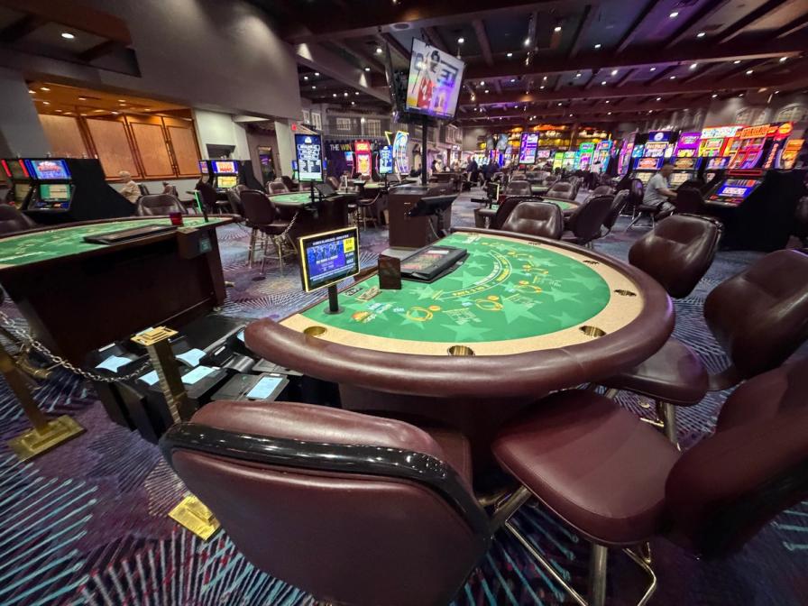 A casino floor featuring a green felt blackjack table with brown leather chairs, and slot machines in the background.