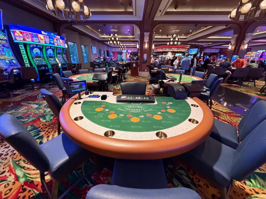 A green felt blackjack table surrounded by blue chairs on a casino floor with slot machines and people in the background.