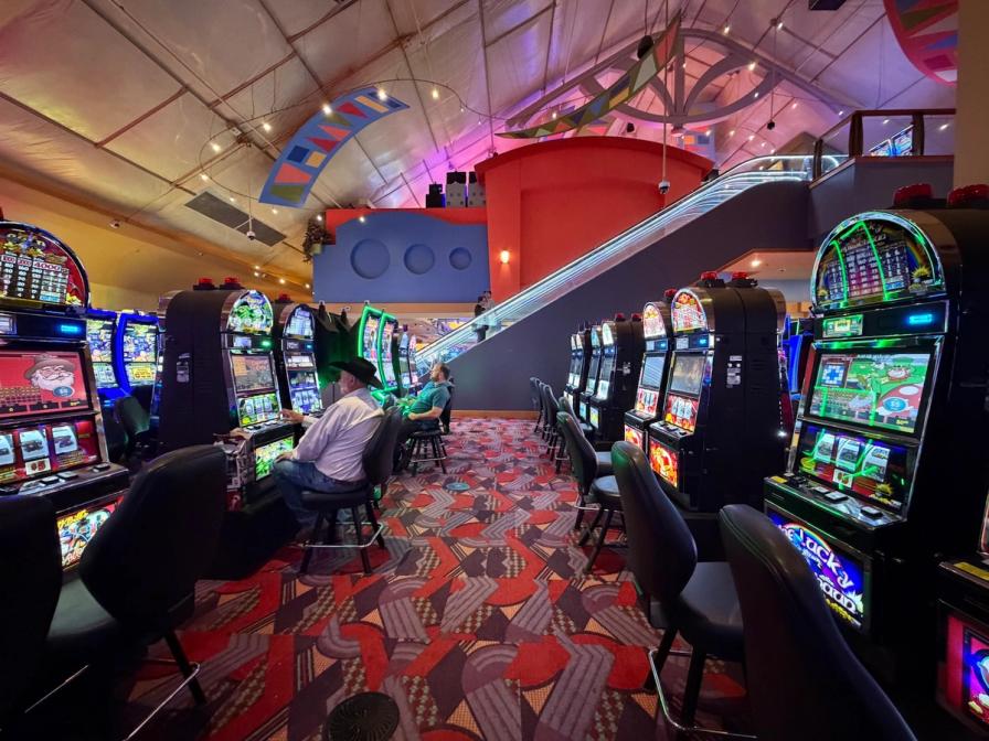 Rows of slot machines in a casino with patterned carpet, an escalator, and a few patrons playing.
