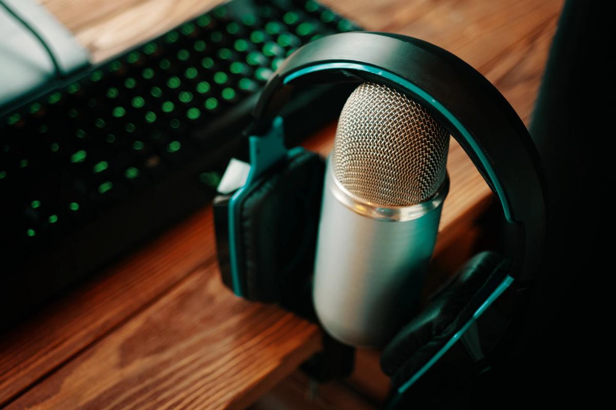 A silver microphone with headphones resting on it, next to a green backlit keyboard on a wooden desk.