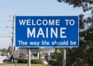 A blue road sign reading "WELCOME TO MAINE The way life should be" against a clear blue sky.