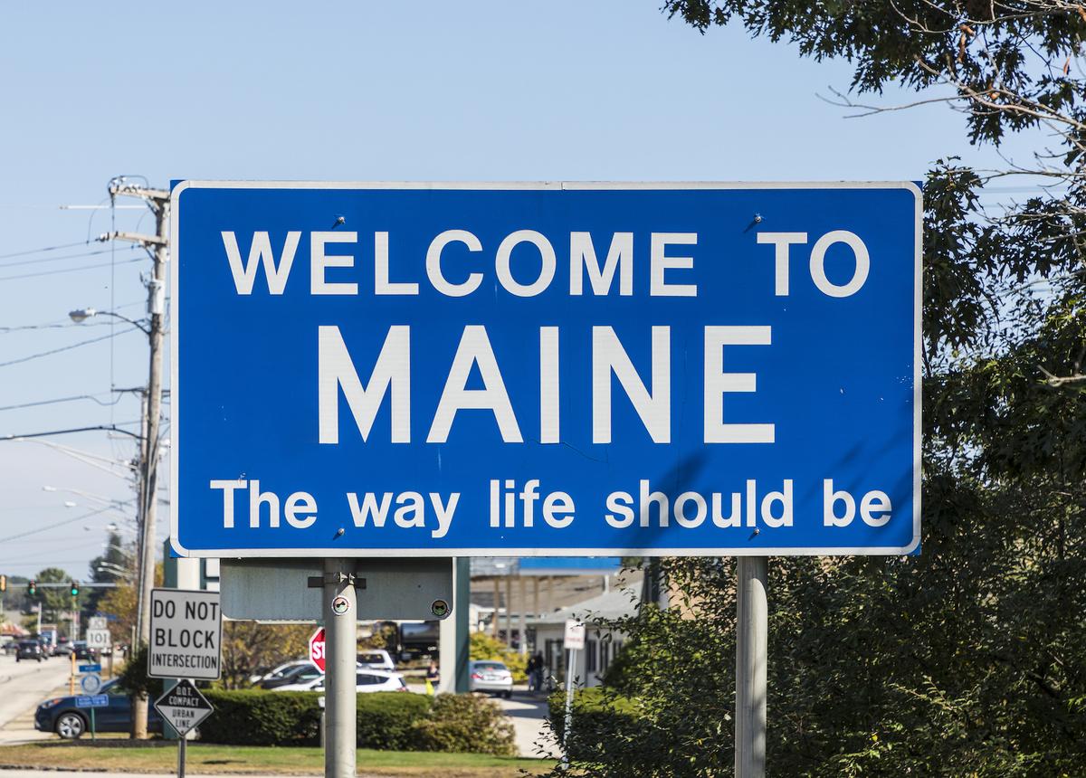 A blue road sign reading "WELCOME TO MAINE The way life should be" against a clear blue sky.