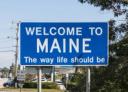 A blue road sign reading "WELCOME TO MAINE The way life should be" against a clear blue sky.