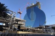Guitar-shaped blue glass hotel under construction in Las Vegas, with red cranes and steel framing against a clear blue sky.