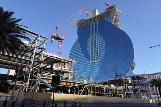 Guitar-shaped blue glass hotel under construction in Las Vegas, with red cranes and steel framing against a clear blue sky.