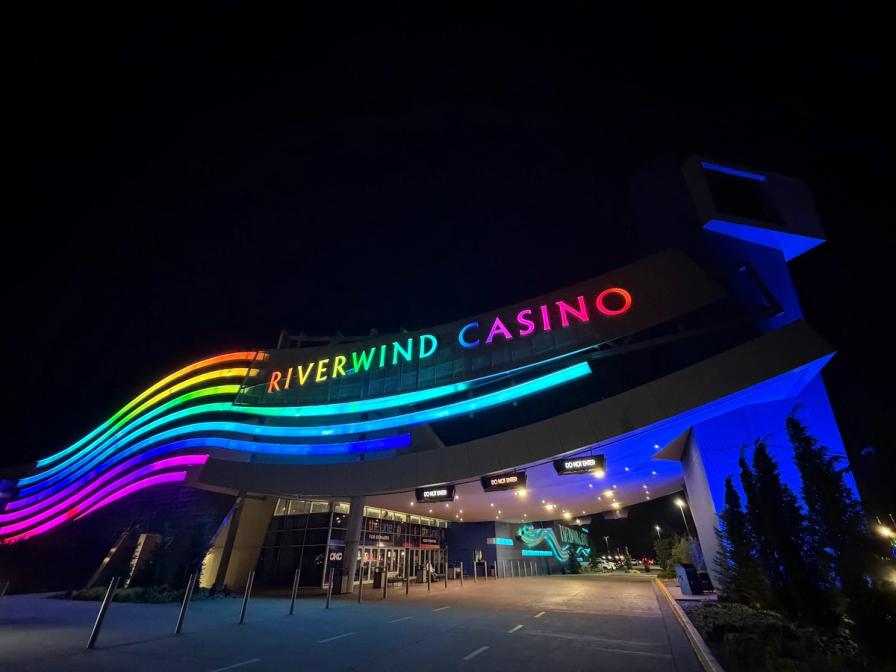 Vibrant rainbow neon sign glows above the entrance of the Riverwind Casino at night under blue architectural lighting.