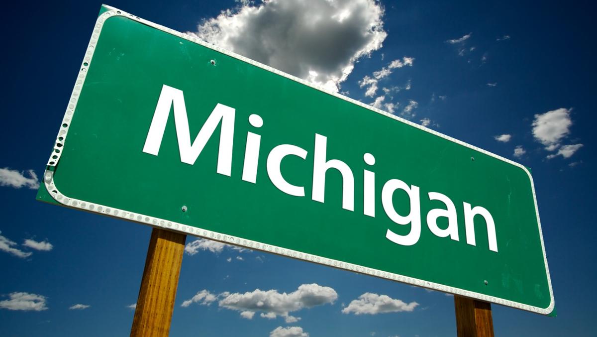 A green highway sign reading "Michigan" in white text, set against a blue sky with clouds.