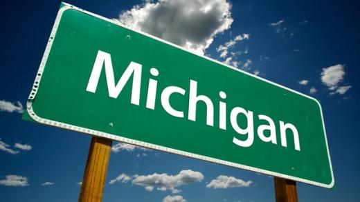 A green highway sign reading "Michigan" in white text, set against a blue sky with clouds.