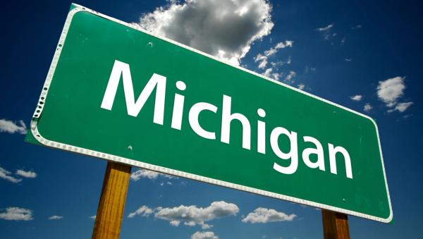 A green highway sign reading "Michigan" in white text, set against a blue sky with clouds.