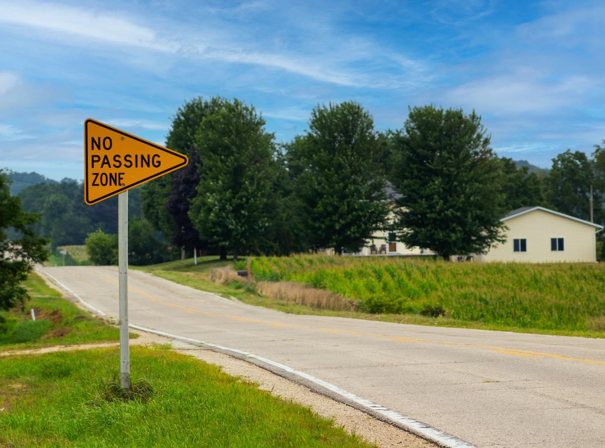 Yellow "No Passing Zone" road sign next to a rural road with trees and a house under a blue sky.