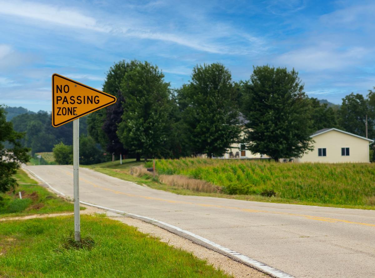 Yellow "No Passing Zone" road sign next to a rural road with trees and a house under a blue sky.