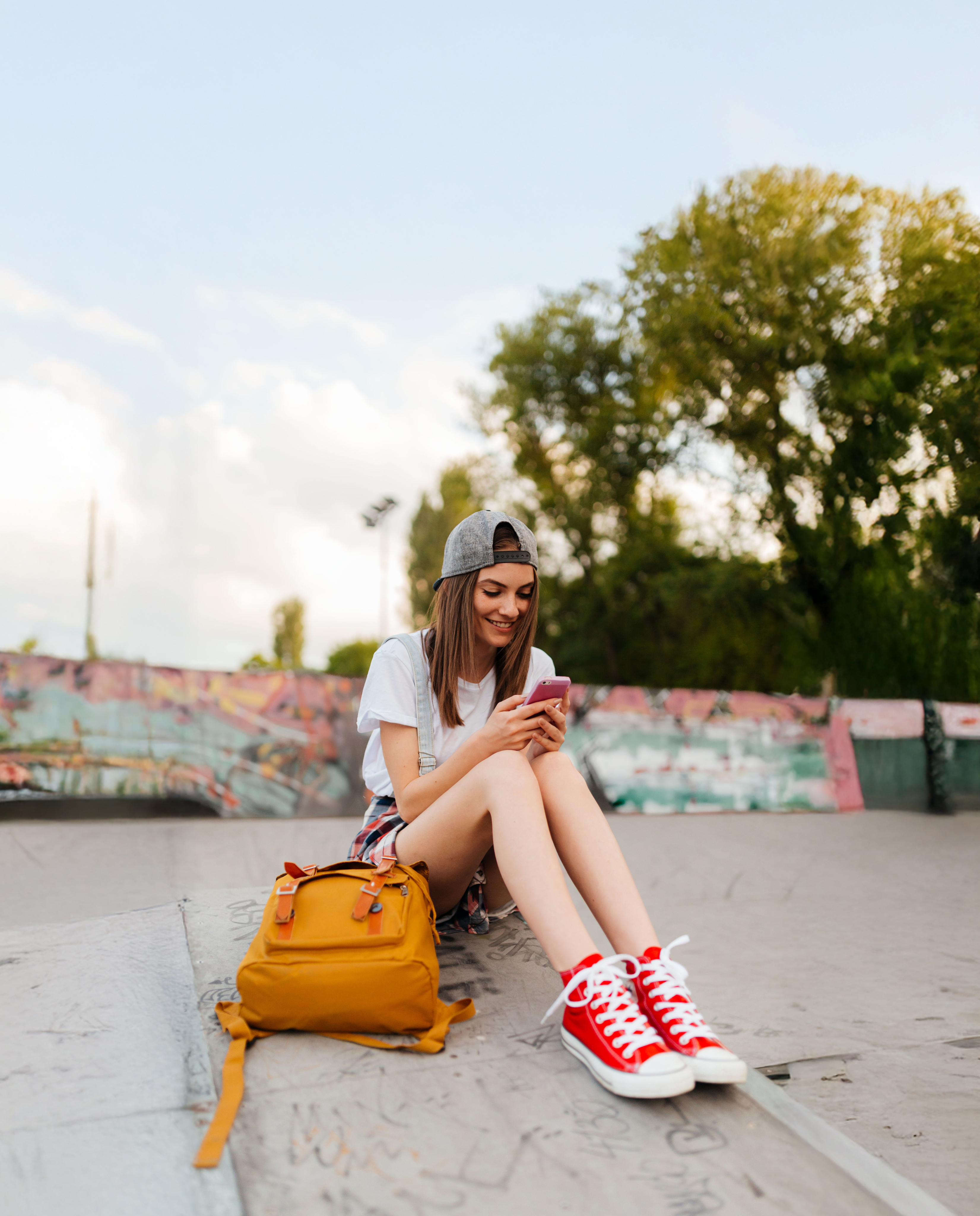 A young woman sits smiling in a skate park, looking at her smartphone, with a yellow backpack beside her.