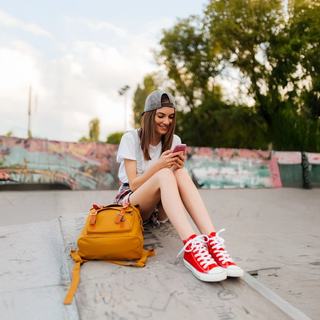 A young woman sits smiling in a skate park, looking at her smartphone, with a yellow backpack beside her.
