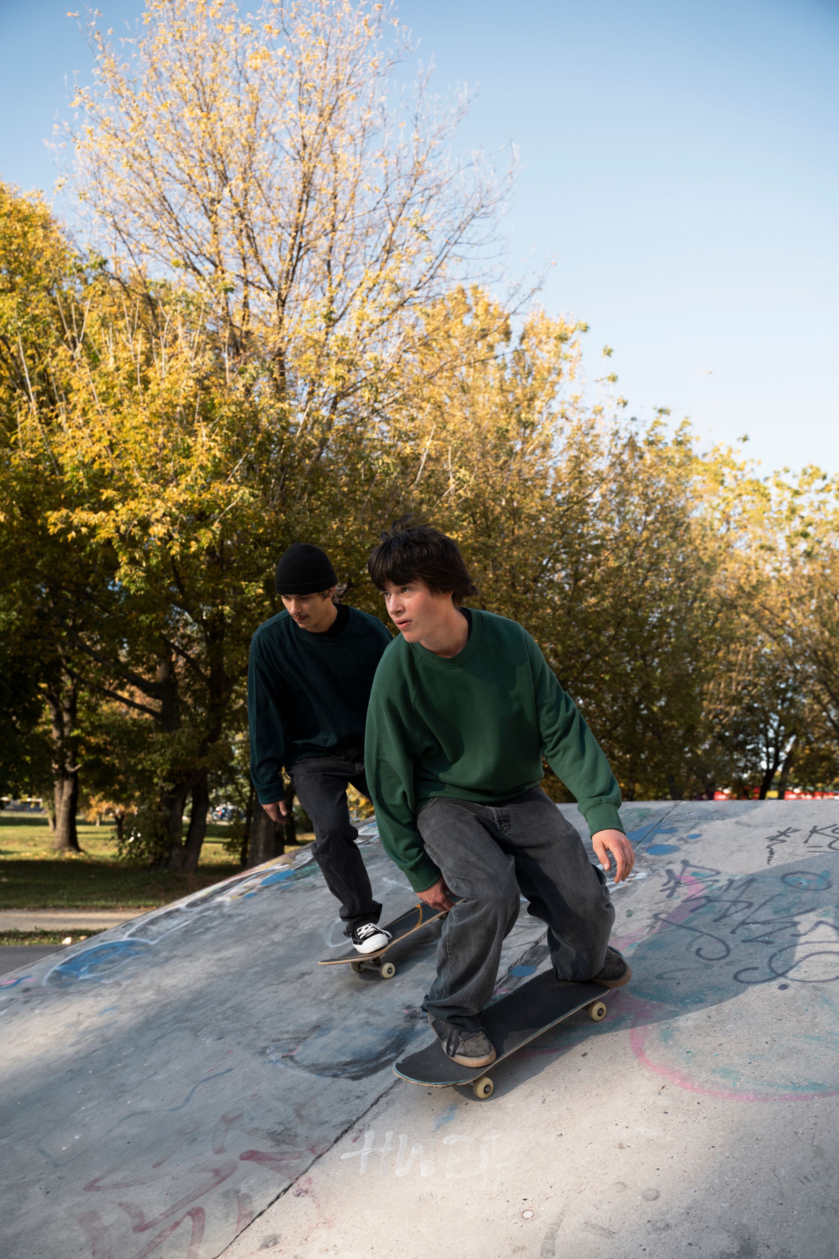 Dos jóvenes patinando en una rampa de concreto, rodeados de árboles con hojas amarillas de otoño.