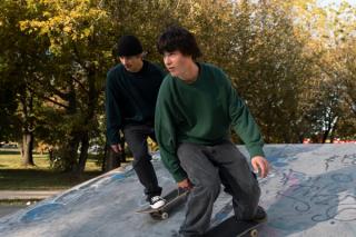 Two young men skateboarding down a concrete ramp, surrounded by trees with yellow autumn leaves.