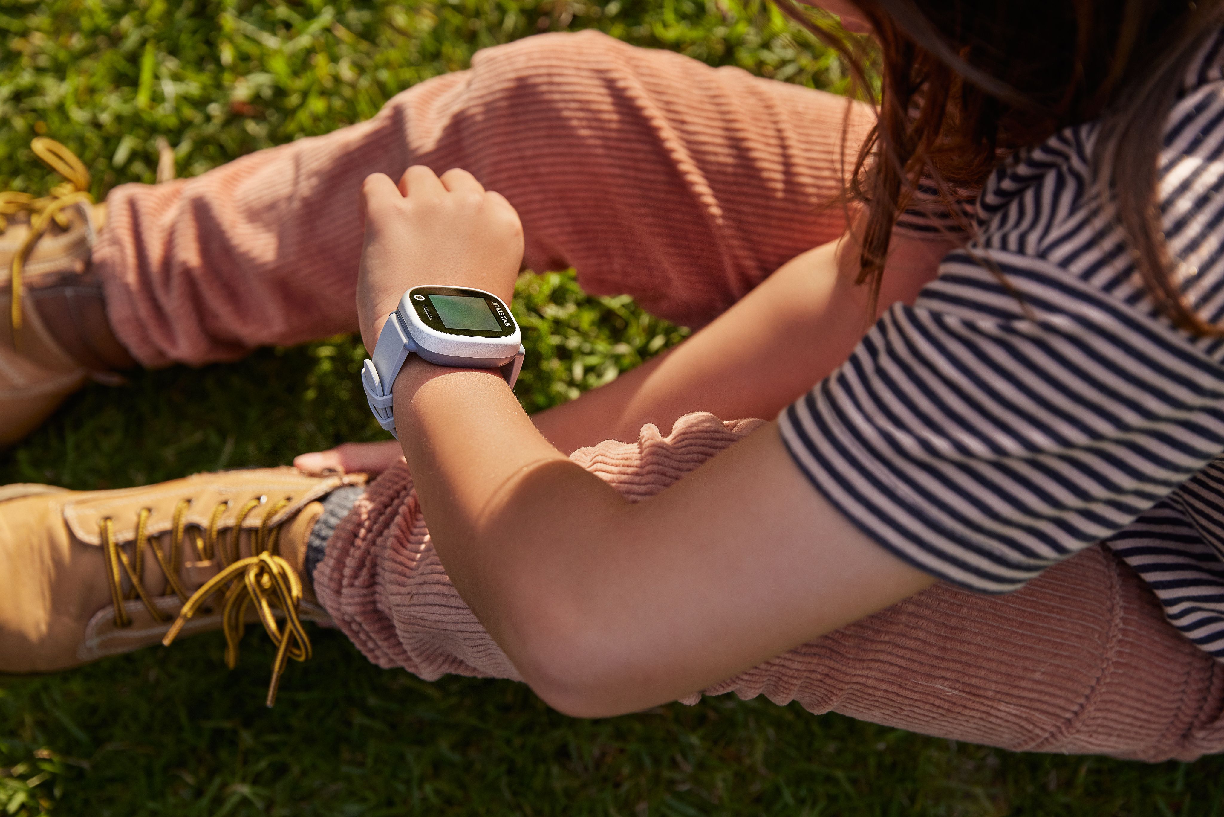 Un niño está sentado en la hierba, mirando un reloj inteligente blanco y azul claro en su muñeca.