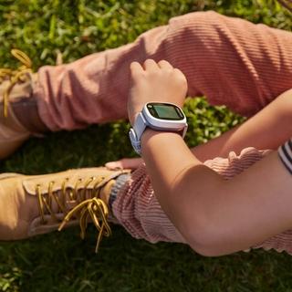 A child sits on grass, looking at a white and light blue smartwatch on their wrist.