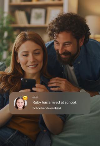 A couple smiles while looking at a smartphone displaying a notification: "Ruby has arrived at school. School mode enabled."