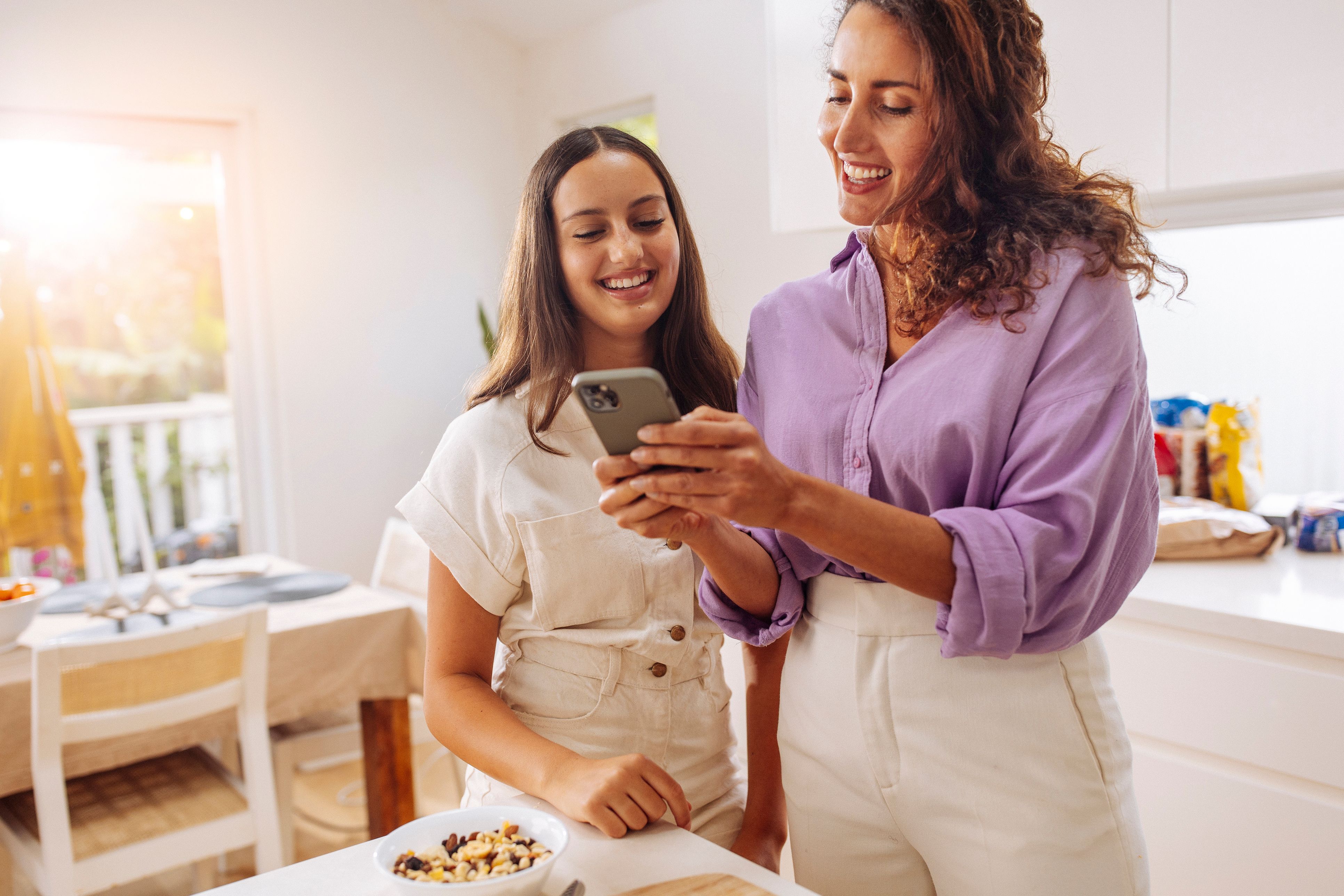 Dos mujeres sonrientes mirando juntas un teléfono inteligente en una cocina luminosa.