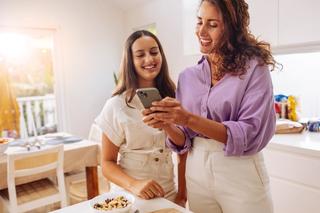Two smiling women looking at a smartphone together in a bright kitchen.