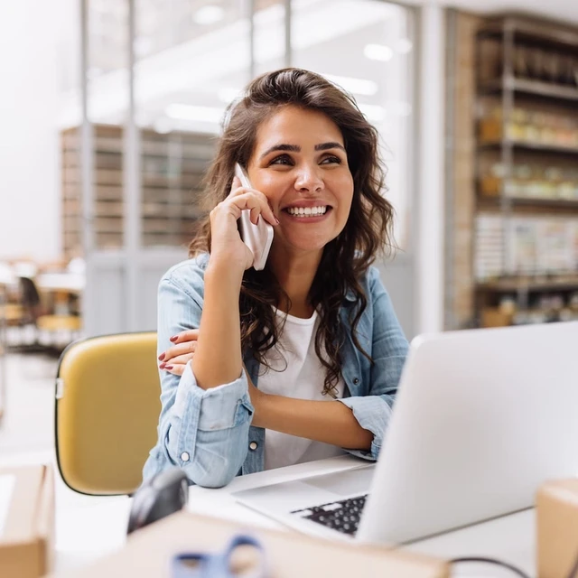 Two warehouse employees, a man and a woman, discussing inventory while holding a clipboard in a storage area filled with labeled cardboard boxes.