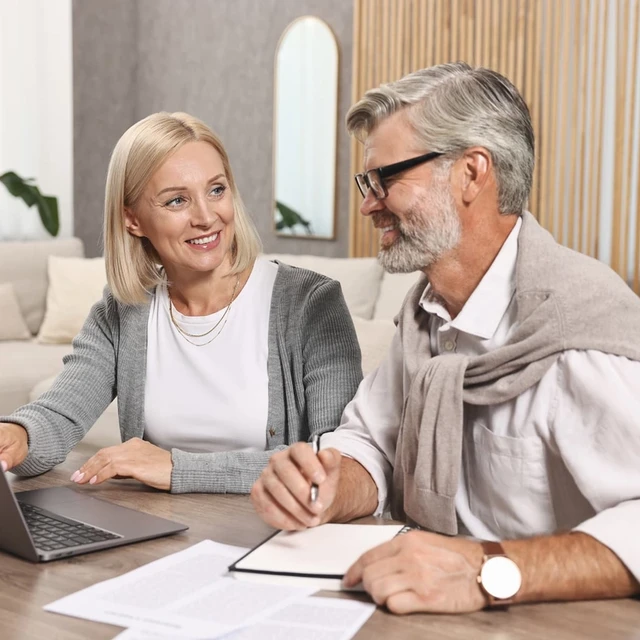 A man working from home, sitting at a desk with a laptop and a dog on his lap, while speaking on the phone. A bookshelf and office supplies are in the background.