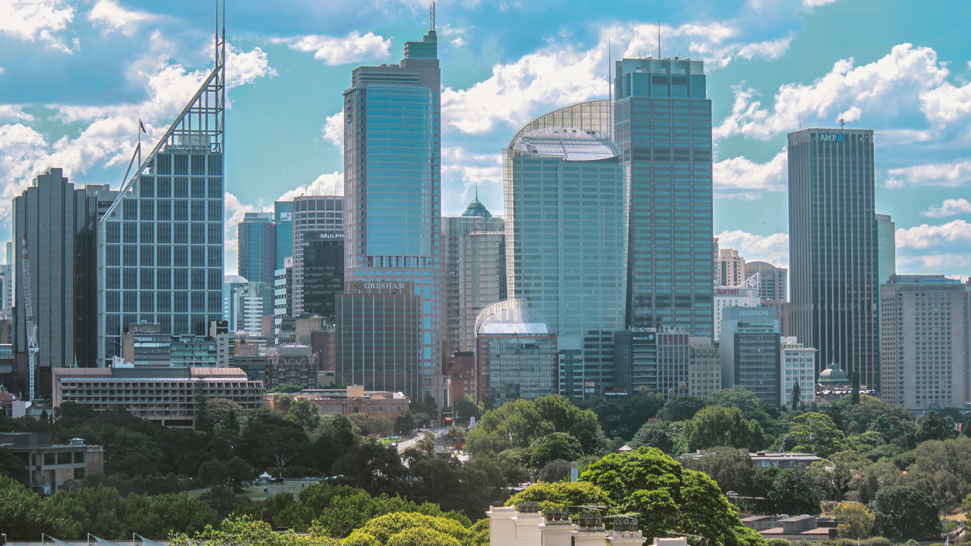 A vibrant cityscape showcasing modern skyscrapers and lush greenery under a bright blue sky with scattered clouds.
