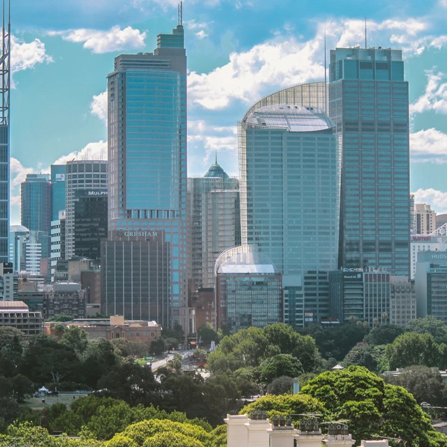 A vibrant cityscape showcasing modern skyscrapers and lush greenery under a bright blue sky with scattered clouds.