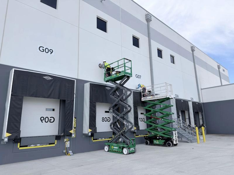 Two installers on scissor lifts applying large dock door number graphics to the exterior wall of a distribution center