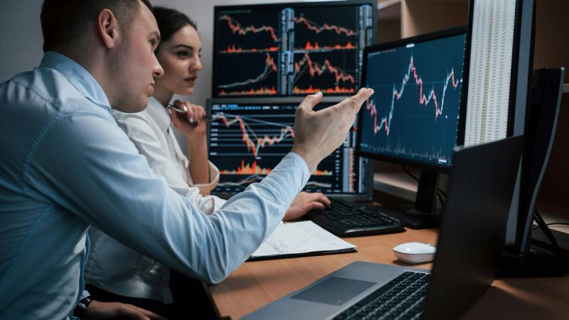 Zwei Trader an einem Multi-Monitor-Arbeitsplatz. Einer erklärt der anderen den Markt am Bildschirm — Begleitung am Touchpoint, eine Stufe vor dem Trade.