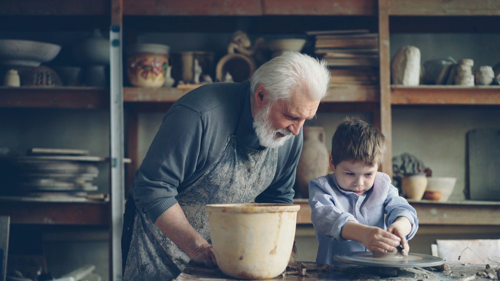 An older man in a work apron leans over a potter's wheel in a workshop, watching a boy shape the clay