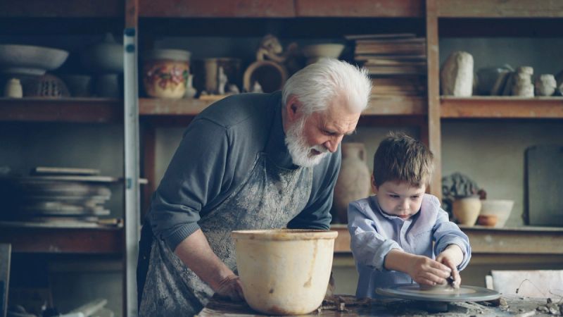 Ein älterer Mann in Arbeitsschürze beugt sich in einer Töpferwerkstatt über die Töpferscheibe, an der ein Junge arbeitet