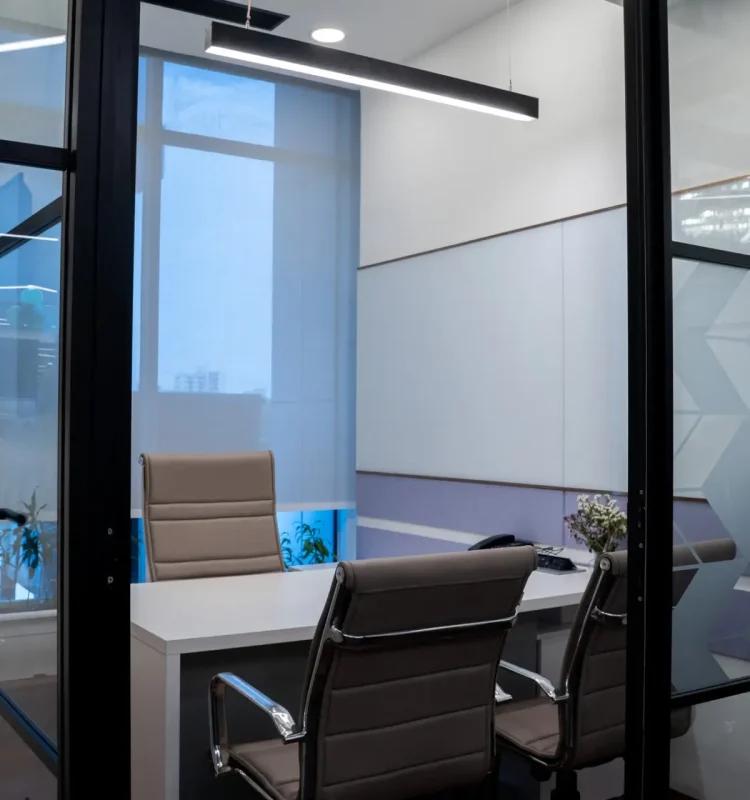 Glass-walled meeting room with a small desk and two guest chairs, seen through an open door