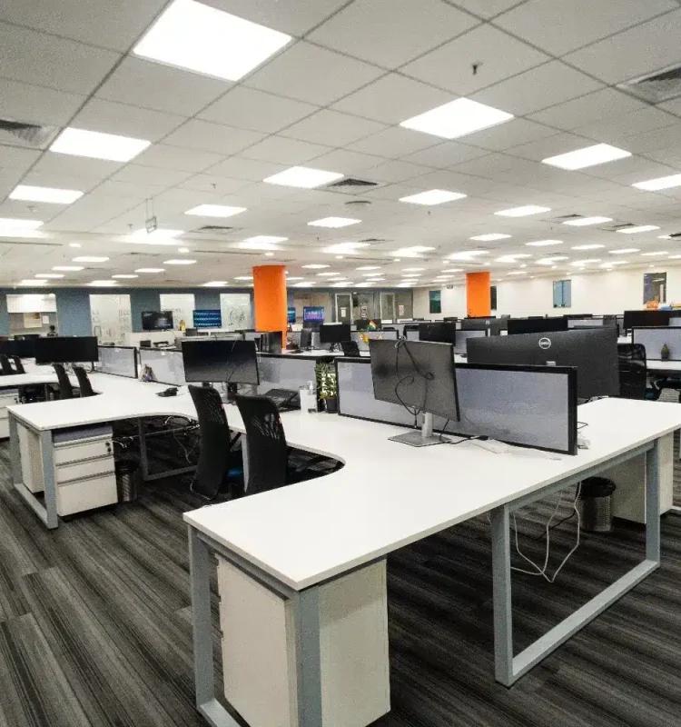 Open-plan workstations with white L-desks, dual monitors and orange pillars under panel lighting, AIHP Signature, Udyog Vihar Gurgaon