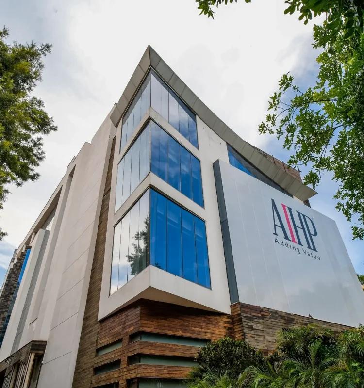 Angular faceted corner glazing and stone-clad base under leafy trees, AIHP Broadway, Sector 32 Gurgaon