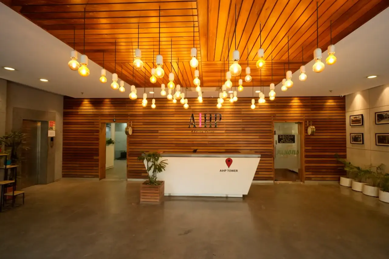Reception under a wood-slat ceiling with hanging pendant bulbs and the AIHP logo, in an AIHP managed office in Gurgaon