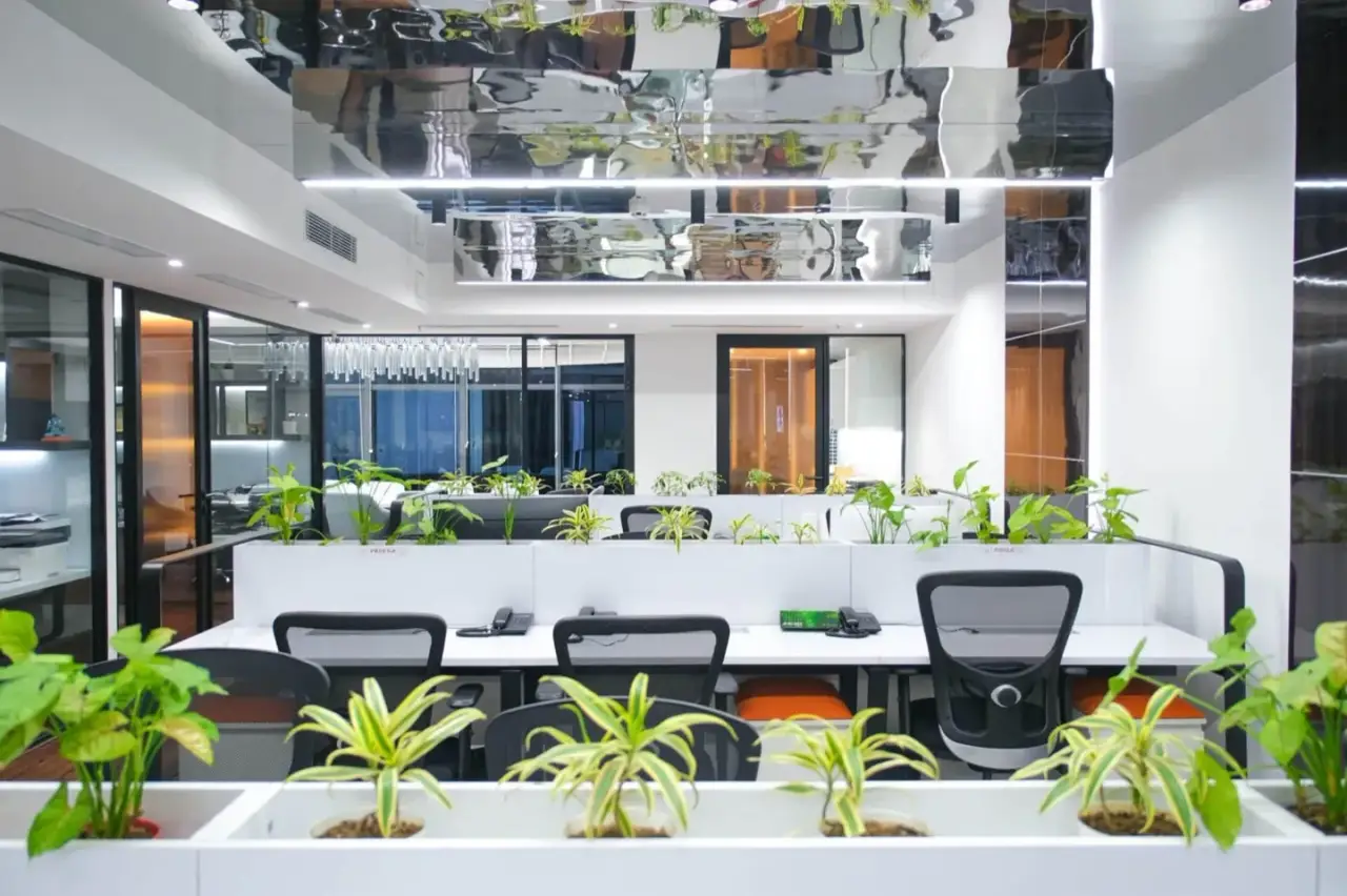 Cluster of white workstations with planter dividers and glass-walled cabins under a mirrored ceiling, in an AIHP managed office in Gurgaon