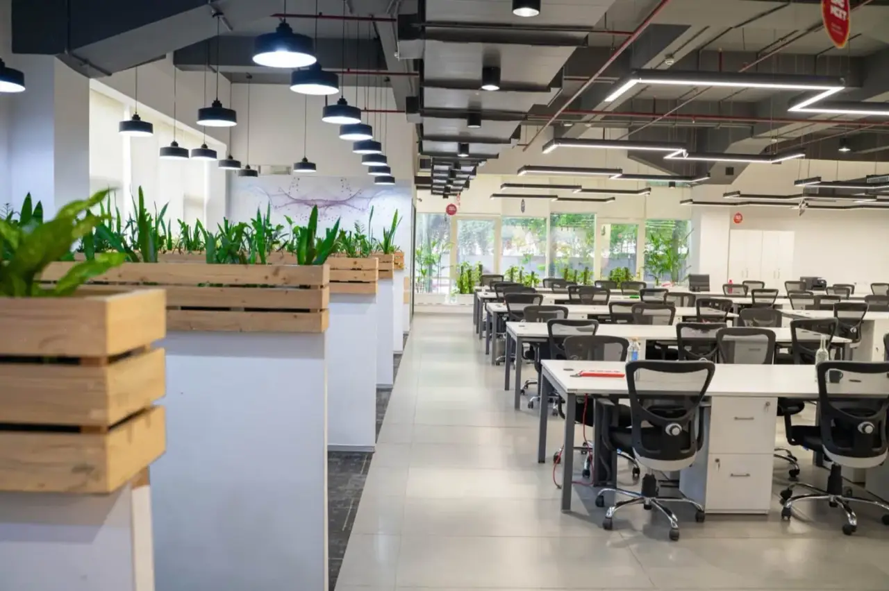 Open-plan workstation floor with an exposed ceiling, pendant lights and wooden planter boxes along the aisle, in an AIHP managed office in Gurgaon