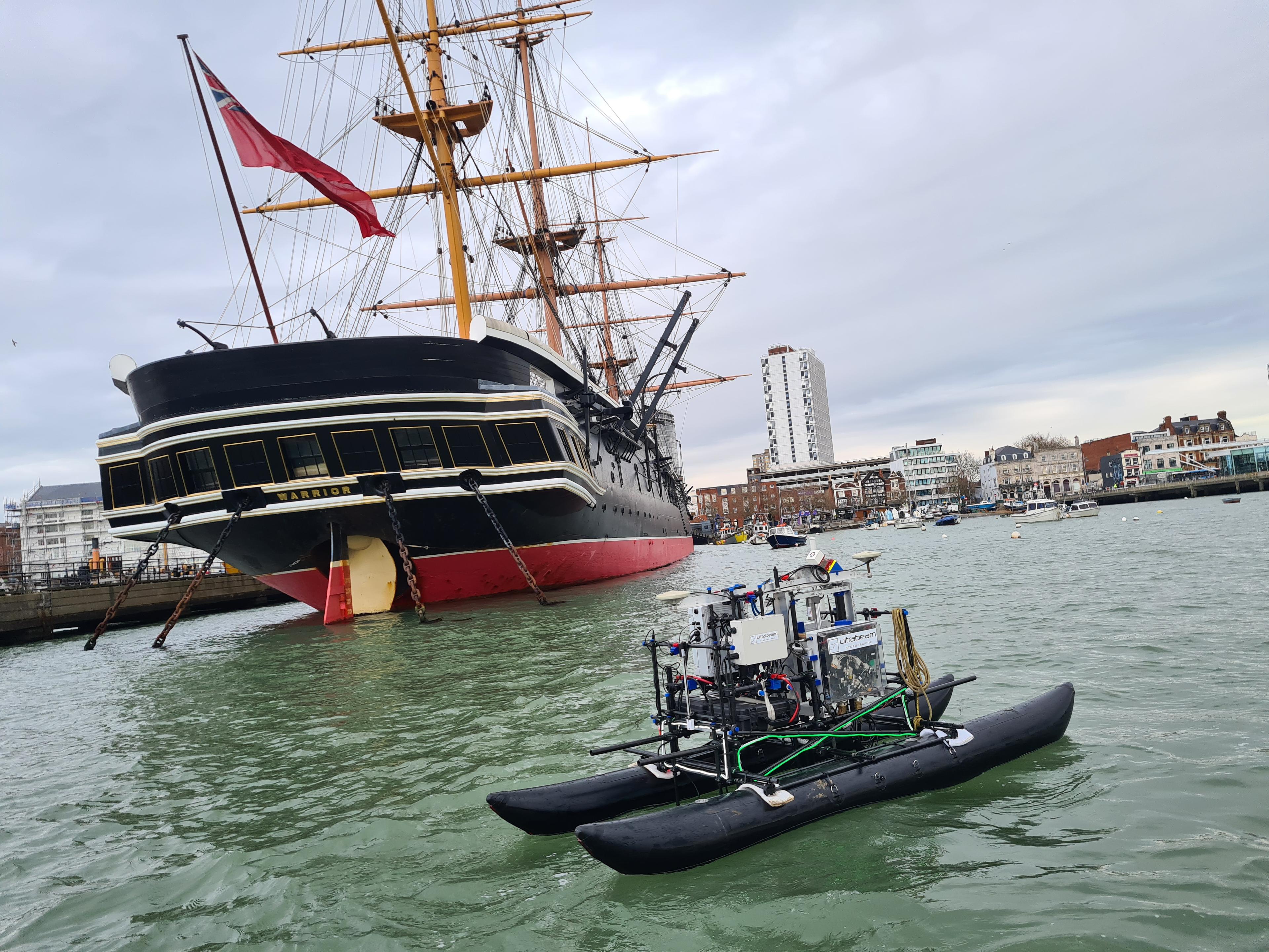 A black autonomous survey vessel on the water in front of a historic sailing ship