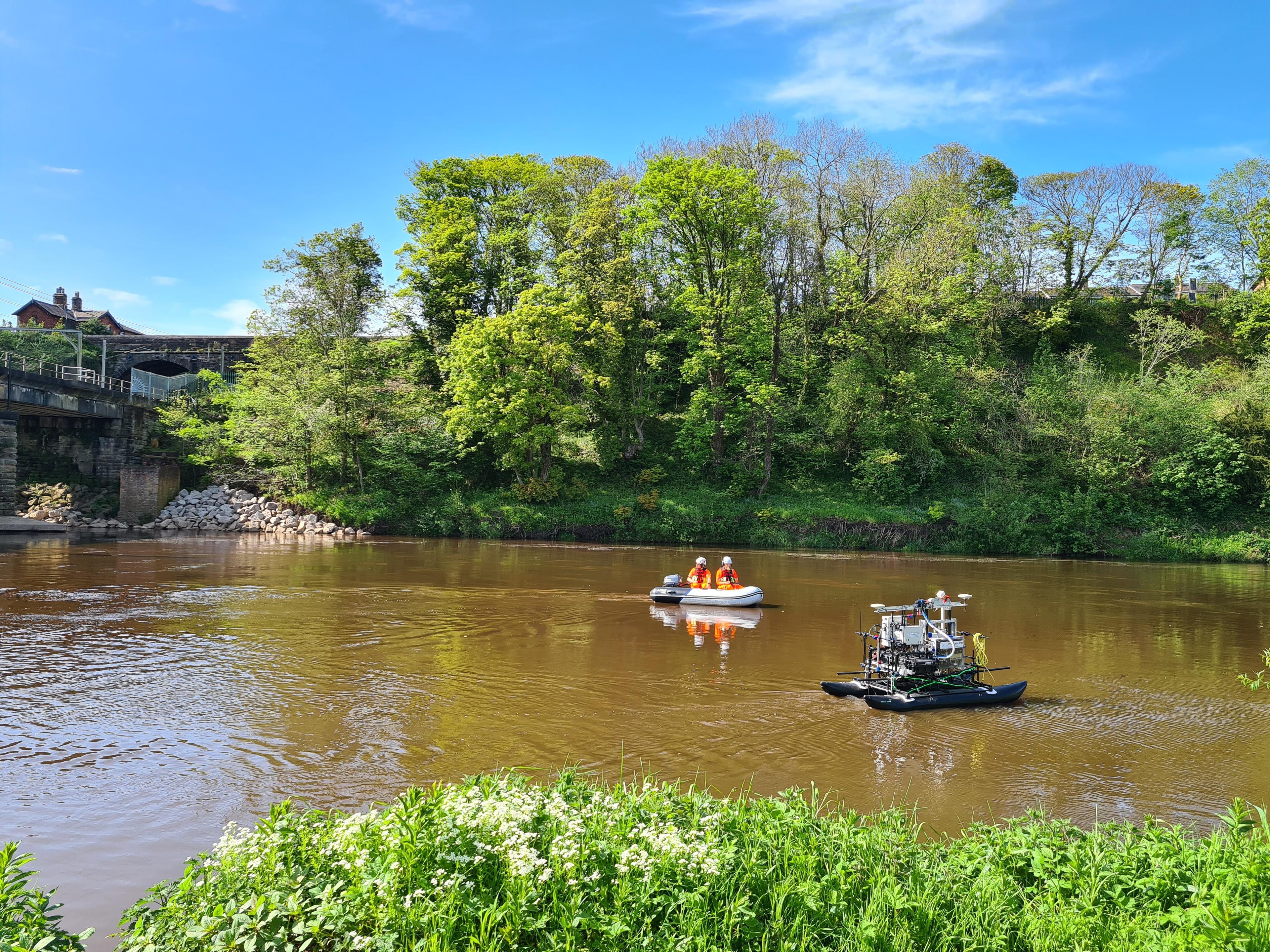 Two men in a boat on a muddy river watch over a autonomous uncrewed vessel carrying out bathymetric survey operations