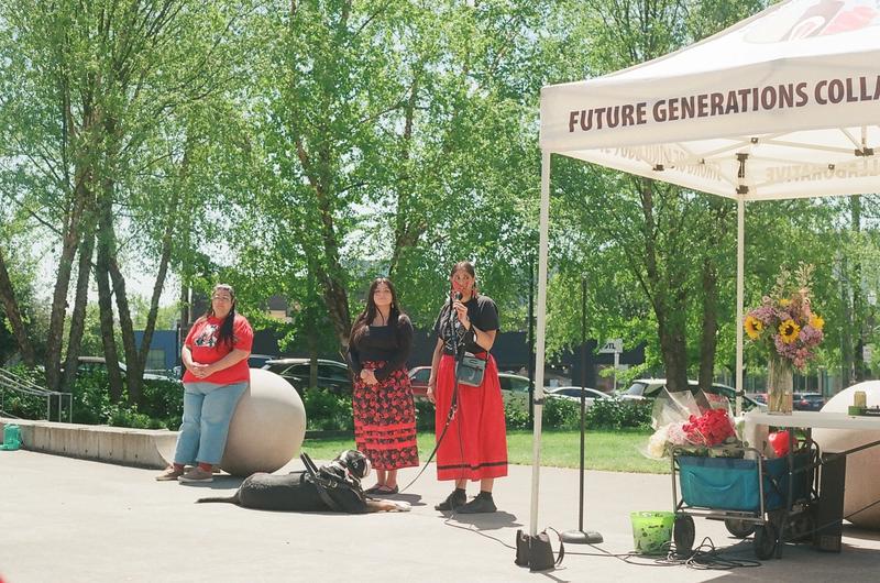 Three women, two in ribbon skirts, stand beside a Future Generations Collaborative tent at an outdoor gathering.