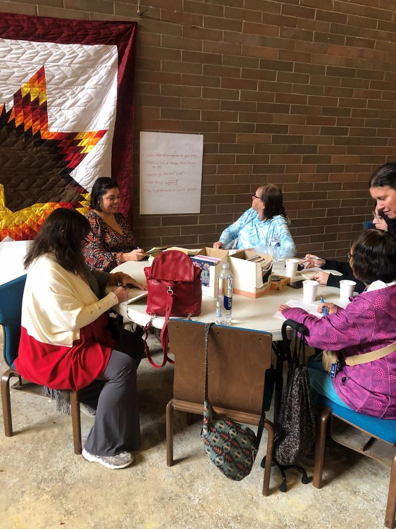 Women sit around a long table working on a craft project, with a star quilt hanging on the brick wall behind them.