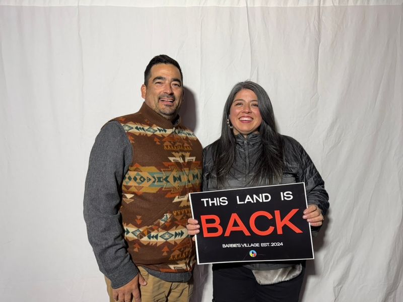 Two community members smile holding a sign that reads “This land is back — Barbie’s Village est. 2024.”