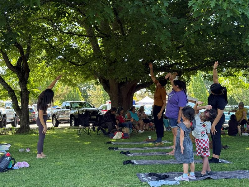 Parents and young children stretch together on yoga mats under large shade trees in a park.